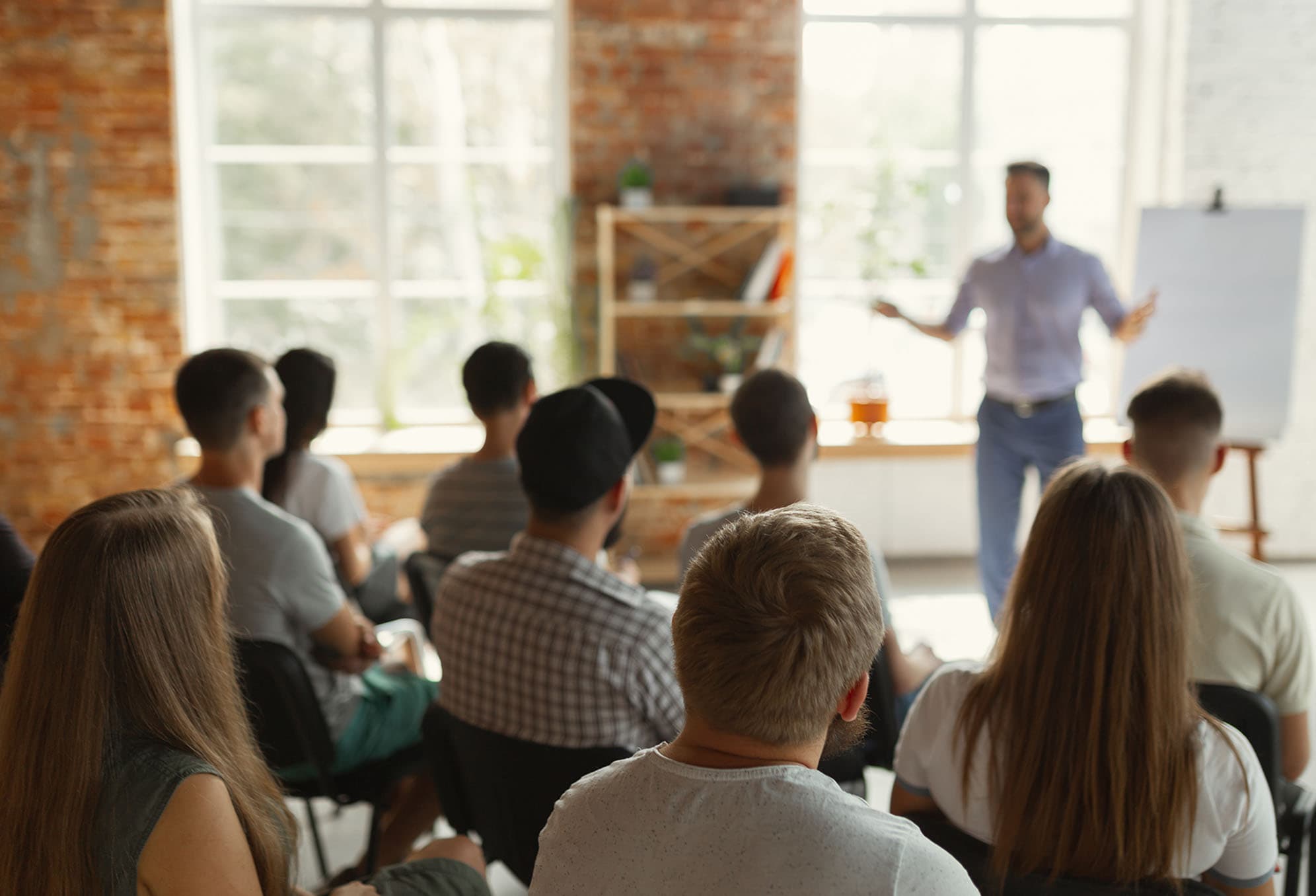 A classroom setting with large windows, and rows of people from the back who are listening to a man standing in front of a white board and easel talking.