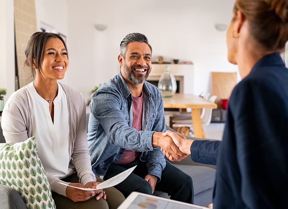 A smiling woman and man seated on a couch. The man is shaking hands with a woman in a blue jacket holding an iPad with a graph on it.